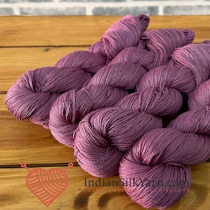 Puede incluir: Varias madejas de hilo color malva profundo dispuestas en un grupo. El hilo tiene una textura suave y ligeramente brillante, retorcido en bucles. El texto IndianSilkYarn.com está impreso en negro cerca del centro inferior de la imagen.