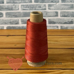 Könnte beinhalten: Eine Spule aus roter Seide steht auf einer Holzoberfläche vor einem Ziegelhintergrund. Der Faden ist eng um einen Pappkern gewickelt. Die Website-Adresse indiansilkyarn.com ist auf der Basis der Spule zu sehen.