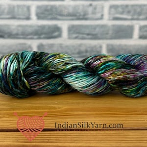 May include: A twisted skein of variegated yarn featuring a blend of deep teal, emerald green, royal purple, and subtle black tones. The yarn has a lustrous, smooth texture. The text IndianSilkYarn.com is printed in black near the bottom.