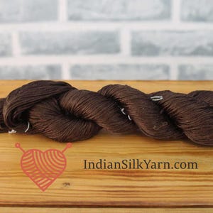 Puede incluir: Un ovillo de hilo marrón oscuro, probablemente de seda, se muestra sobre una superficie de madera. El hilo está torcido y parece suave. La imagen incluye un logotipo con un diseño en forma de corazón y el texto "IndianSilkYarn.com".