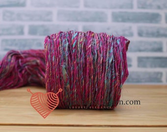May include: A vibrant skein of yarn in shades of pink, teal, and yellow. The yarn is tightly wound into a rectangular shape, with loose strands visible. The background features a brick wall and a wooden surface. The image includes a logo with the text "Luxury, Elegance, Beauty."