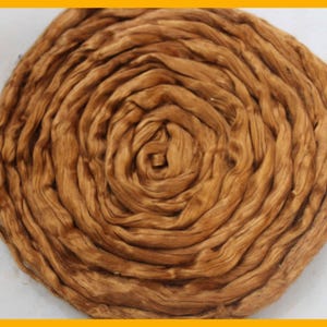 May include: A close-up of a tightly wound bundle of golden-brown fibres, possibly silk or wool. The fibres are arranged in a spiral pattern, creating a circular design. The texture appears soft and slightly lustrous, with subtle variations in colour.