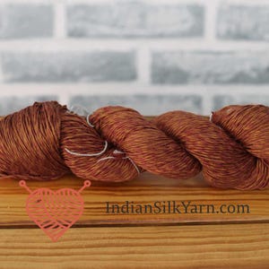 Puede incluir: Primer plano de tres madejas de hilo marrón con reflejos rojos. El hilo está sobre una superficie de madera, con un fondo de ladrillos. El texto "IndianSilkYarn.com" es visible.