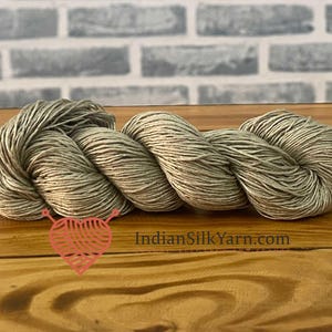 May include: A skein of light green yarn, tightly wound, rests on a wooden surface. The yarn is a neutral color, with a slight sheen. The background features a light gray brick wall. The text "IndianSilkYarn.com" is visible.