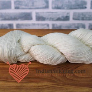 以下が含まれることがあります： 木製の表面に、白っぽい色の糸が束ねられ、流れるような形に配置されています。糸の質感は自然光によって強調されています。「IndianSilkYarn.com」のテキストとハート型のロゴが見えます。