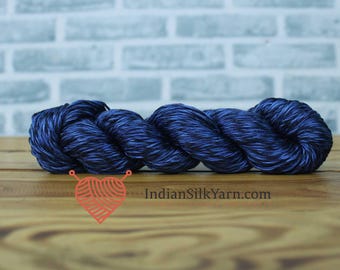 May include: A skein of dark blue yarn, tightly twisted, with subtle variations in color. The yarn is displayed on a wooden surface against a light blue brick wall. The text "IndianSilkYarn.com" is visible.