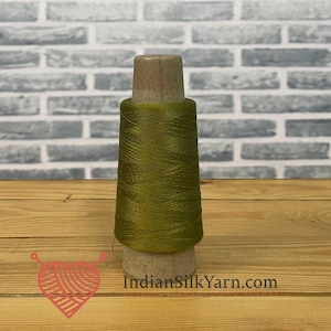 May include: A spool of olive green silk yarn. The yarn is tightly wound around a wooden core. The background is a light gray brick wall and a wooden surface. The website address "IndianSilkYarn.com" is visible.
