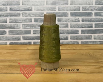 May include: A spool of olive green silk yarn. The yarn is tightly wound around a wooden core. The background is a light gray brick wall and a wooden surface. The website address "IndianSilkYarn.com" is visible.