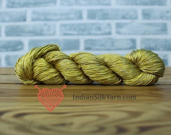 May include: A skein of golden-yellow yarn, tightly wound and textured, rests on a wooden surface. The yarn appears to be silk, with subtle variations in color. The website IndianSilkYarn.com is visible in the image. Ideal for knitting and crochet projects.