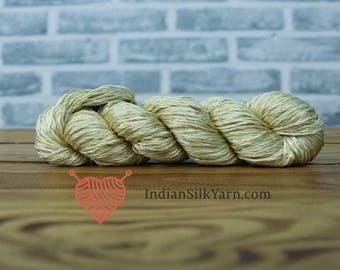 May include: A skein of natural-colored silk yarn, tightly wound and ready for crafting. The yarn has a subtle sheen and a soft, inviting texture. The background features a light blue brick wall and a wooden surface. The logo "IndianSilkYarn.com" is visible.