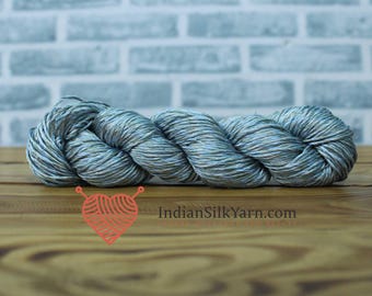May include: A skein of blue and gray yarn, ideal for knitting or crochet projects, rests on a wooden surface. The yarn is tightly wound, showing a blend of colors. The background features a light blue brick wall. The website IndianSilkYarn.com is visible.
