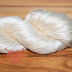 Peut inclure: Deux écheveaux de fil de soie blanc cassé sont présentés sur une surface en bois. Le fil est étroitement enroulé, avec une texture lisse et brillante. L'image comprend un logo en forme de cœur et le texte "IndianSilkYarn.com".