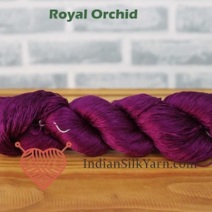 Puede incluir: Un ovillo de hilo color Royal Orchid. El hilo es de un tono púrpura profundo y rico, con un ligero brillo. El texto "Royal Orchid" se muestra encima del hilo. El sitio web "IndianSilkYarn.com" se muestra debajo.