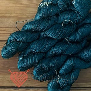 Può includere: Una collezione di matasse di filato color verde ottanio profondo con una finitura lucida e brillante. Il filato è attorcigliato in più fasci, mostrando un ricco colore blu-verde scuro. Un piccolo logo arancione a forma di cuore è visibile.