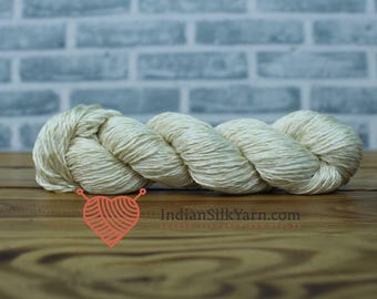 May include: A skein of off-white silk yarn, tightly wound, rests on a wooden surface. The yarn has a soft, natural sheen. The background features a light blue brick wall. The logo "IndianSilkYarn.com" is visible.
