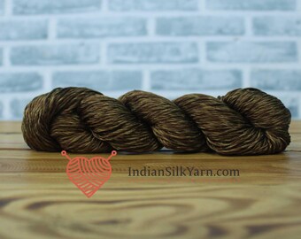 May include: A skein of brown yarn, likely silk, is displayed on a wooden surface. The yarn has a rich, earthy tone with subtle variations in color. The background features a light blue brick wall. The website address "IndianSilkYarn.com" is visible.