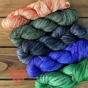 May include: A collection of six skeins of yarn arranged in a diagonal stack. The colors include peach, olive green, dark charcoal, deep blue, bright royal blue, and vibrant emerald green. Each skein features a twisted, multi-tonal fiber texture.