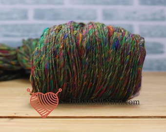 May include: A close-up of a colorful yarn ball. The yarn is a mix of green, brown, red, and yellow threads. The yarn is on a wooden surface with a light blue brick background. The image includes a logo that says "IndianSilkYarn.com".