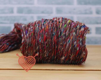 May include: A close-up of a vibrant, multi-colored yarn skein. The yarn is a mix of red, brown, and other bright colors. The yarn is tightly wound and has a textured appearance. The image also includes a logo with the text "IndianSilkYarn.com".
