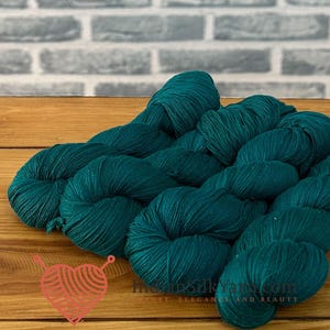 Op de afbeelding: Verschillende strengen diepblauwgroen garen zijn in een groep gerangschikt. Het garen heeft een gladde, licht glanzende textuur. Een klein rood hartvormig logo met breinaalden en de tekst "IndianSilkYarn.com" is zichtbaar in de linkerbenedenhoek.
