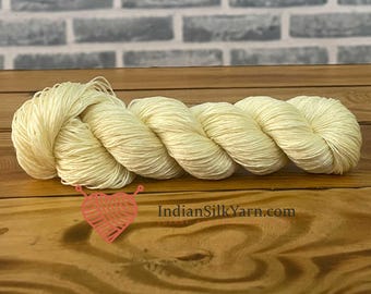 May include: A skein of ivory-colored yarn, tightly wound and displayed on a wooden surface. The yarn is arranged in a series of loops, showcasing its texture and form. The website address IndianSilkYarn.com is visible.