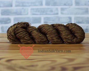 May include: A skein of brown and gold yarn, ideal for knitting or crochet projects. The yarn is tightly wound and has a textured appearance. The background is a light brick wall and a wooden surface. The text "IndianSilkYarn.com" is visible.