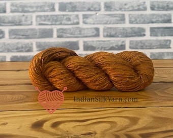 May include: A skein of orange-brown yarn, tightly wound, rests on a wooden surface. The yarn has a textured appearance, with visible strands. The background features a light gray brick wall. The website IndianSilkYarn.com is visible.