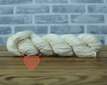 May include: A skein of off-white yarn, tightly wound, resting on a wooden surface. The yarn is a creamy color, with visible texture. The background features a light blue brick wall. The text "IndianSilkYarn.com" is visible.