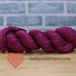 May include: A skein of vibrant magenta yarn, tightly wound, rests on a wooden surface. The yarn is a rich, saturated color, with visible texture and sheen. The background features a light blue brick wall. The text "IndianSilkYarn.com" is visible.