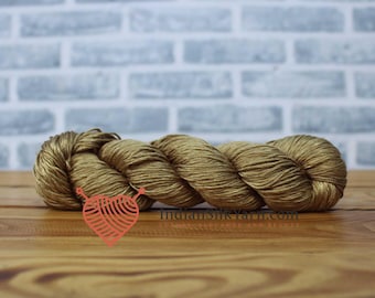May include: A skein of golden-brown yarn, tightly wound, rests on a wooden surface. The yarn has a subtle sheen, suggesting a silk or similar fiber. The background features a light gray brick wall. The logo of IndianSilkYarn.com is visible.