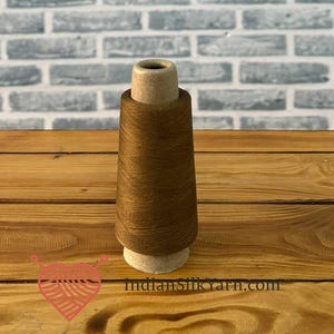 Può includere: Una bobina di filo marrone è appoggiata su una superficie di legno. Il filo è avvolto attorno a un'anima di cartone. Lo sfondo presenta un muro di mattoni grigio chiaro. La bobina è di un colore marrone caldo, ideale per vari progetti di artigianato.