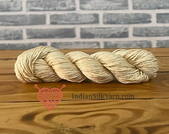 May include: A skein of natural-colored yarn, likely silk, is displayed on a wooden surface. The yarn is twisted into a compact bundle, showcasing its texture and sheen. The background features a brick wall. The image includes a logo and the text "IndianSilkYarn.com".