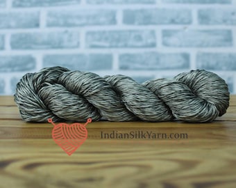 May include: A skein of gray and white yarn, ideal for knitting or crochet projects. The yarn is tightly wound and appears soft, with a subtle sheen. The background features a brick wall and a wooden surface.