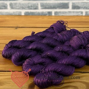 Puede incluir: Una colección de madejas de hilo de color morado intenso con un brillo distintivo, dispuestas en grupo. El hilo presenta una textura suave y retorcida con un color vibrante y saturado. Un pequeño logotipo con un corazón y agujas es visible.