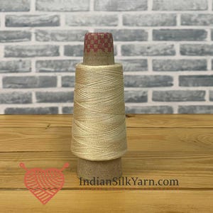 May include: A spool of light beige silk yarn, suitable for crafting. The yarn is wound around a cardboard core, with a red and brown checkered pattern on top. The background features a brick wall and wooden surface. The website address "IndianSilkYarn.com" is visible.