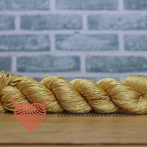 Puede incluir: Un ovillo de hilo de seda amarillo dorado, bien enrollado y expuesto sobre una superficie de madera. El hilo es de un color rico y cálido, con un brillo sutil. El fondo presenta una pared de ladrillos azul claro. El sitio web indiansilkyarn.com es visible.
