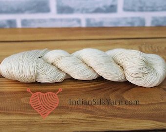 May include: A skein of off-white yarn is displayed on a wooden surface. The yarn is tightly wound and appears soft. The background features a blurred brick wall. A logo with a heart shape and the text "IndianSilkYarn.com" is visible.