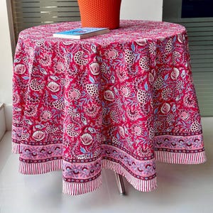 May include: A round, red tablecloth with a floral pattern in white, light blue, and pink. The tablecloth is draped over a small table. A small book and a terracotta pot with a plant are on top of the table.