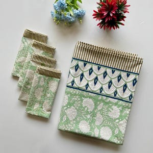 Puede incluir: Un juego de cinco servilletas de algodón verde y blanco con estampado floral y un mantel verde a juego con estampado floral y un borde de rayas marrones, conchas blancas y detalles azules.