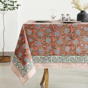 May include: A coral tablecloth with a floral pattern of white and blue flower-like designs and green leaves. The tablecloth has a striped border and is draped over a wooden table.