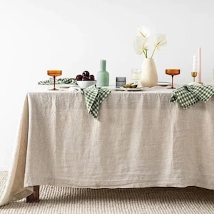 May include: A beige linen tablecloth covers a wooden table set for a meal. The table is decorated with a white vase of flowers, green and white checkered napkins, amber-colored glasses, and a bowl of dark purple fruit.