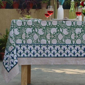 Puede incluir: Un mantel con estampado floral verde y blanco. El mantel tiene un borde con un diseño de abanico azul y blanco y un borde a rayas. La mesa es de madera.