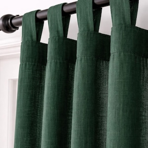 May include: Dark green tab-top curtains hanging from a black curtain pole. The curtains are made of a textured fabric and are partially closed, creating soft folds. The colour is a deep, rich shade of green. Home decor.