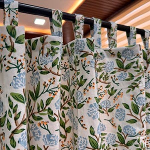 May include: Cream-coloured floral curtains with a repeating pattern of green leaves, blue flowers, and brown branches. The curtains are hanging from a black rod with fabric loops. The design is a botanical theme.