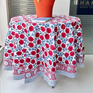 Puede incluir: Un mantel redondo con un estampado floral rojo y azul sobre fondo blanco. El mantel cubre una mesa, con un borde decorativo. Una maceta naranja con una planta y un libro están sobre la mesa.