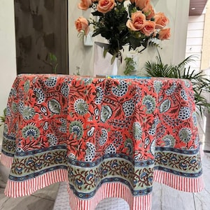 Puede incluir: Un mantel redondo de color coral con un estampado floral en azul, verde y marrón. El borde presenta un diseño a rayas rojas y blancas. Un ramo de rosas melocotón está sobre la mesa.
