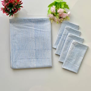 Op de afbeelding: Een opgevouwen wit en blauw gestreept tafelkleed met vier bijpassende servetten. De blauwe strepen zijn verticaal en gelijkmatig verdeeld. De items zijn gerangschikt op een wit oppervlak, met roze en rode bloemen op de achtergrond.