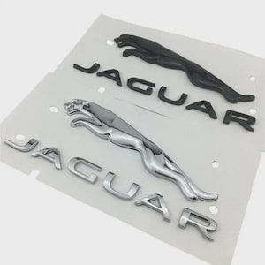 Könnte beinhalten: Zwei Jaguar-Autoembleme. Eines hat eine glänzend schwarze Oberfläche, das andere eine glänzende Chromoberfläche. Beide zeigen das springende Jaguar-Logo und das Wort "JAGUAR" in Großbuchstaben. Die Embleme befinden sich auf weißem Hintergrund.