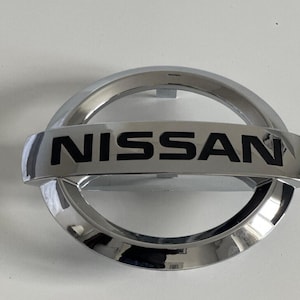 Peut inclure: Un emblème de voiture argenté avec le mot "NISSAN" en lettres noires.