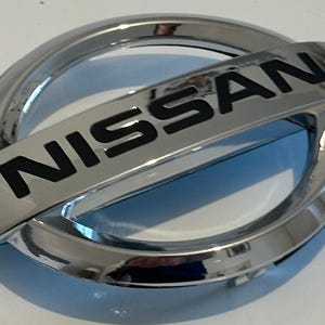 Peut inclure: Un emblème de voiture chromé avec le mot "NISSAN" en lettres noires.